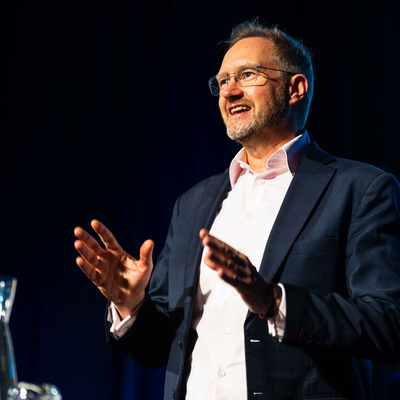 Ben Harris presenting on stage at the 2026 All Actuaries Summit, speaking with both hands raised against a dark backdrop.