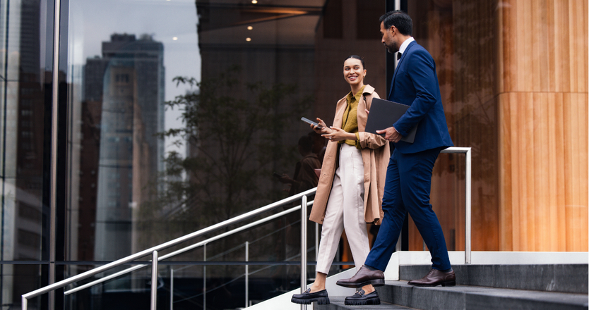 Two business professionals, a woman and a man, walk down exterior stairs beside a glass-fronted building