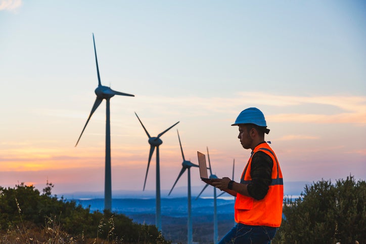 Young electrical engineer business man standing in front of wind turbines checking and working about technical problems and writes the results of measurements with laptop pc in wind power plant electric energy station