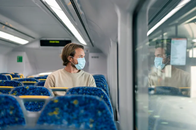 A male passenger sits on an empty train. He is looking out of the window. The Caucasian male is wearing a face mask due to the covid-19 pandemic. The man is commuting to the office as he is an essential worker.