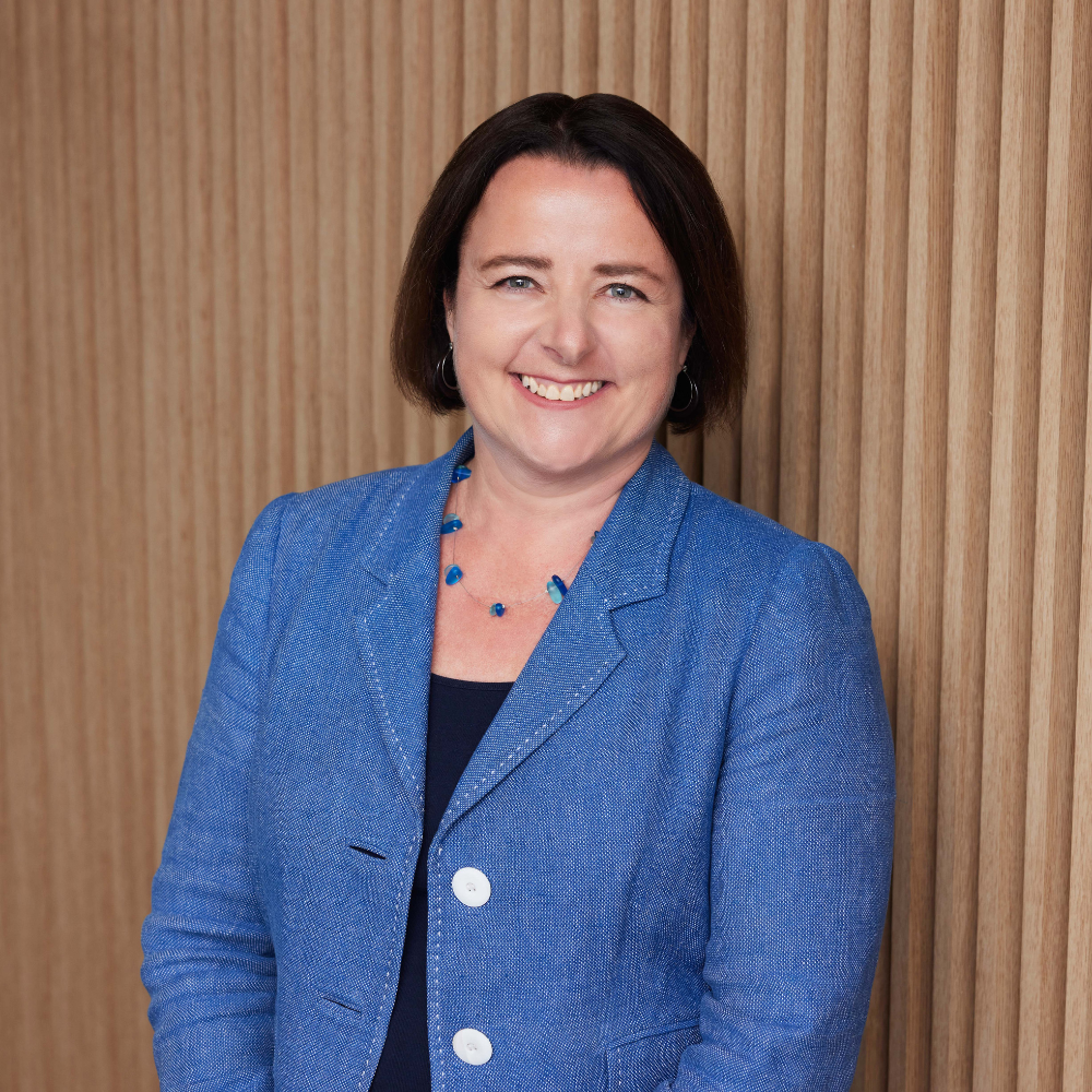 Portrait photo of Elayne Grace smiling to camera - photo from waist up with blue jacket and blue beaded necklace