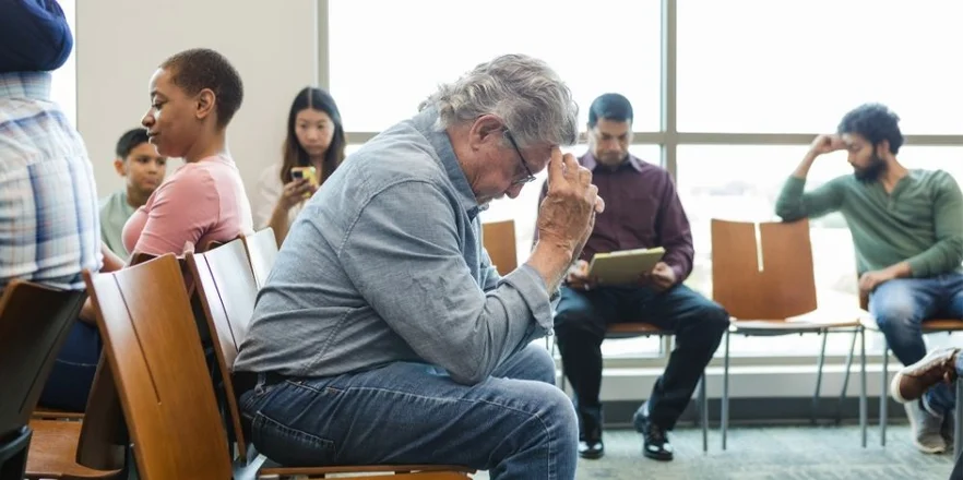 The senior adult man sits anxiously in the waiting room with his hands against his forehead