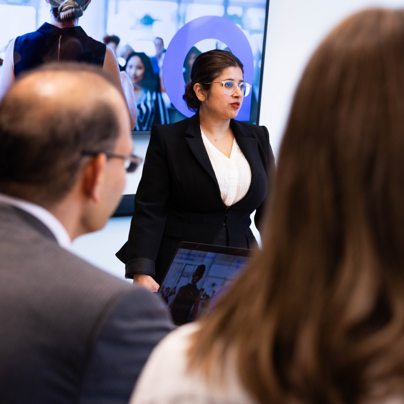 Female standing in a room in front of presentation. Wearing black jacket and white shirt, has dark hair pulled back.