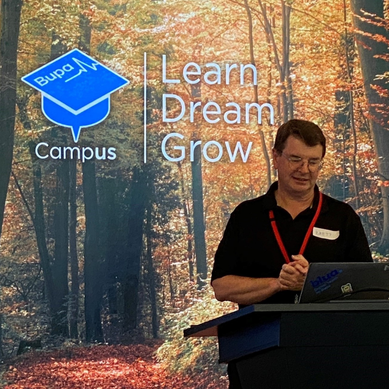 Man speaking at a lectern with black shirt and backdrop of trees and company logo
