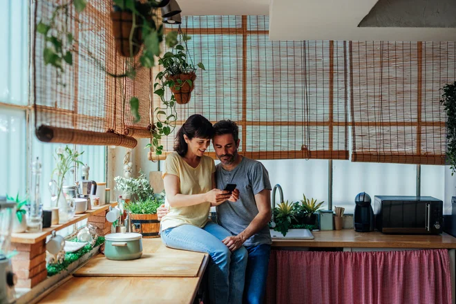 A middle aged Caucasian couple is bonding in the kitchen. The wife is sitting on the countertop showing her husband something on her smartphone