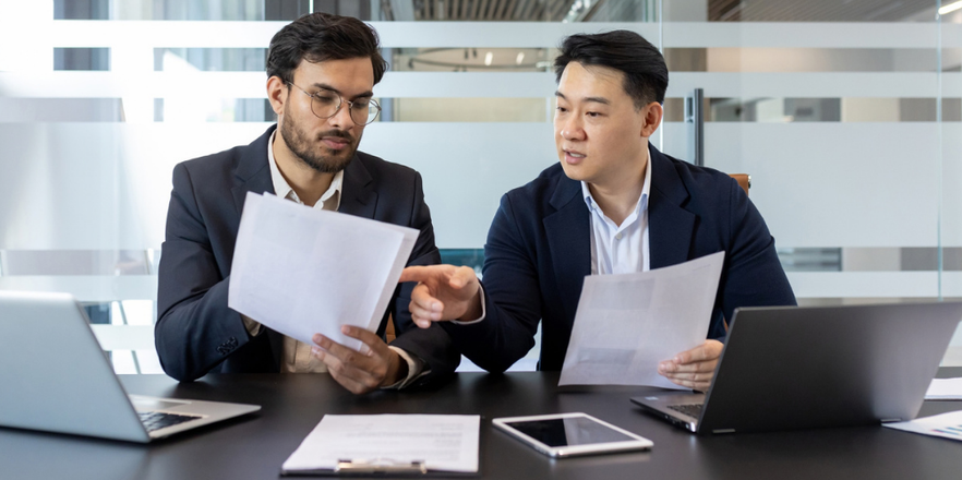 Two men sitting at boardroom table discussing reports.