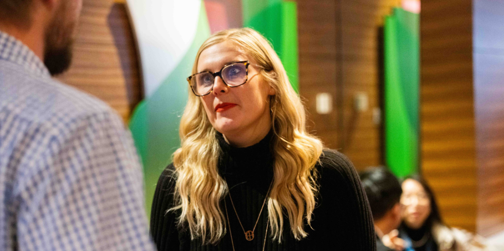 Woman in black polo and long blonde hair, glasses in a work meeting area looking to another person