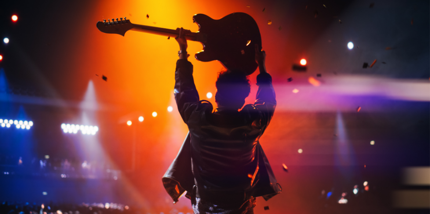 Rock band performing on stage, guitarist in a leather jacket celebrating a successful concert with devoted audience. Footage from the back.