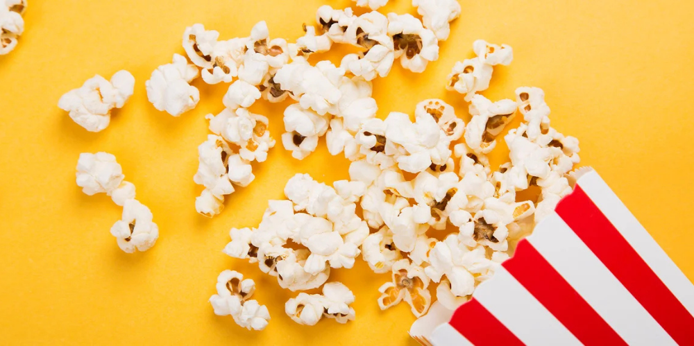 A red and white striped popcorn box spilling popcorn against a bright yellow background.