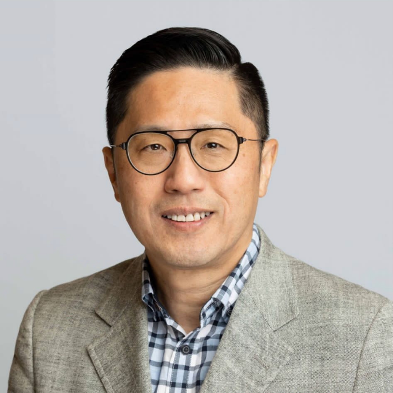 Man with glasses looking at camera and smiling with a light coloured jacket and a blue and white checked shirt