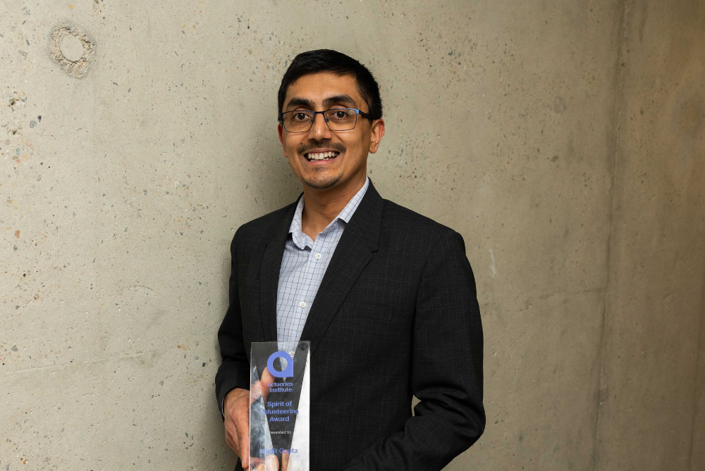 Business man with dark hair and moustache, and open neck blue and white checked shirt, smiling at camera and holding a trophy