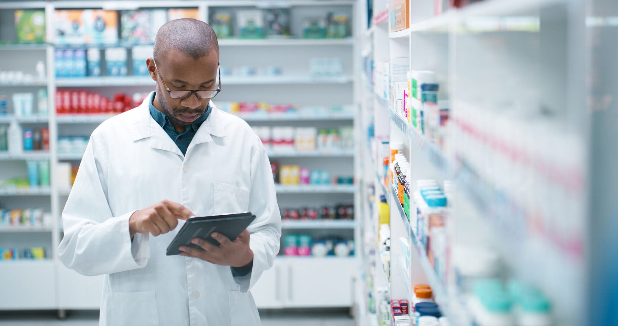 Black man, pharmacist and tablet with shelf at dispensary for checklist, stock or inventory at pharmacy. Young African, male person or medical employee on technology for healthcare or pharmaceuticals