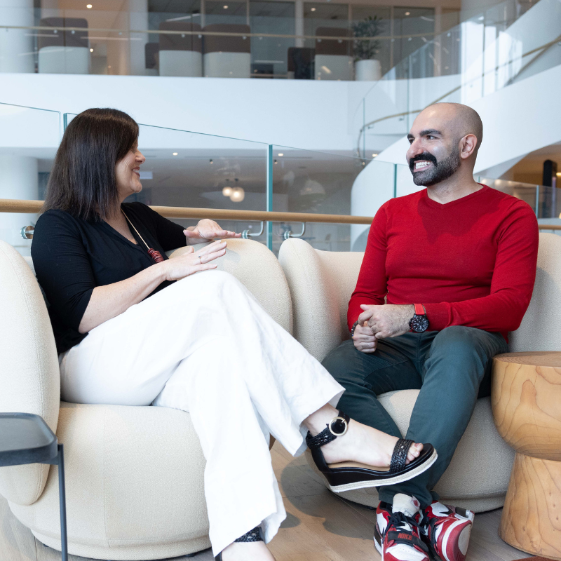 Woman sitting on biege lounge on left, in black pants and white top looking at man who is smiling with grey pants and red long sleeved top