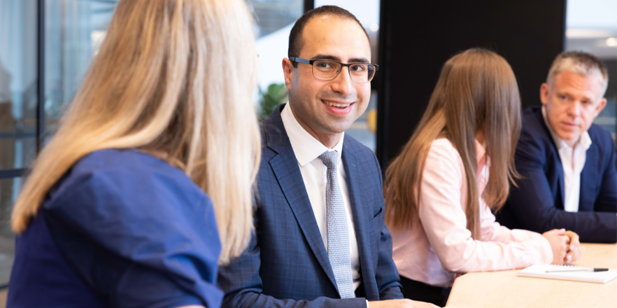Four professionals seated around a meeting table in a modern office. A man in a navy suit and glasses smiles as he engages in conversation with a colleague across the table.