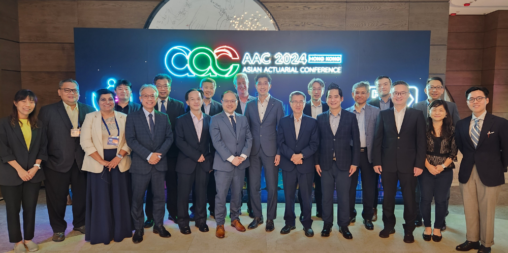 Group of men and women of different cultural backgrounds stand in front of conference sign.
