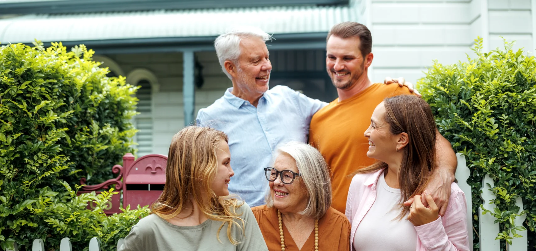 Smiling multigenerational family gathered outside a heritage home with white picket fence and garden hedges.