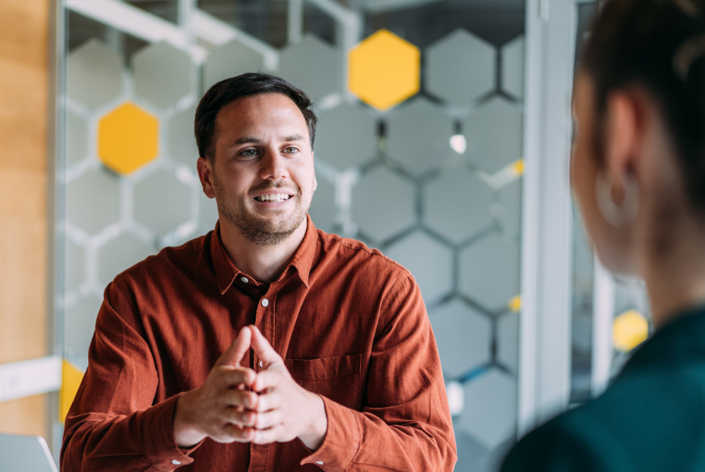 Male sitting at a desk with brown shirt on and frosted glass background with hexagons, talking to a women in dark green shirt with hair in a bun.