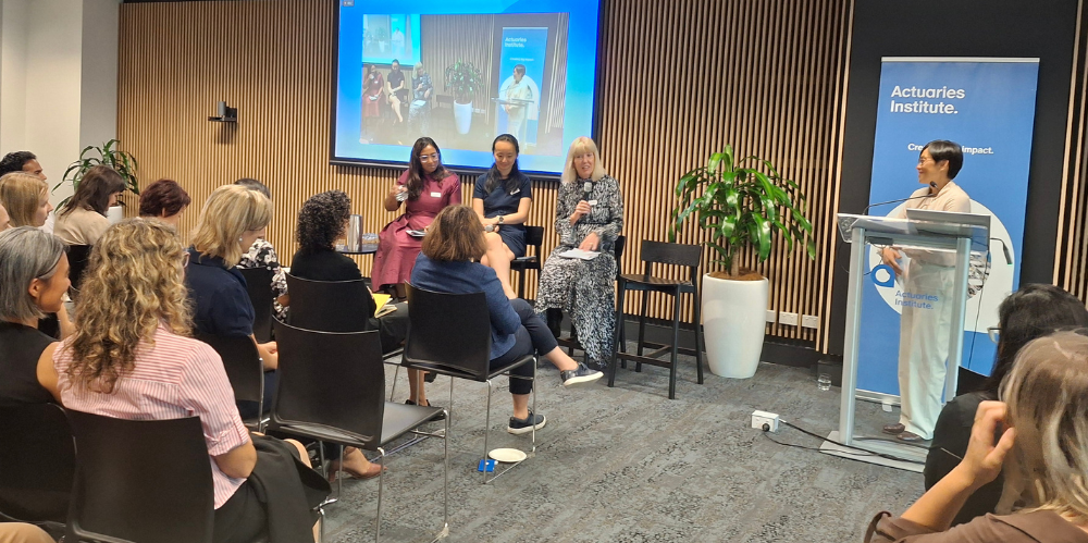 Seminar room of women with hour females up front, three on high stools and one at the microphone
