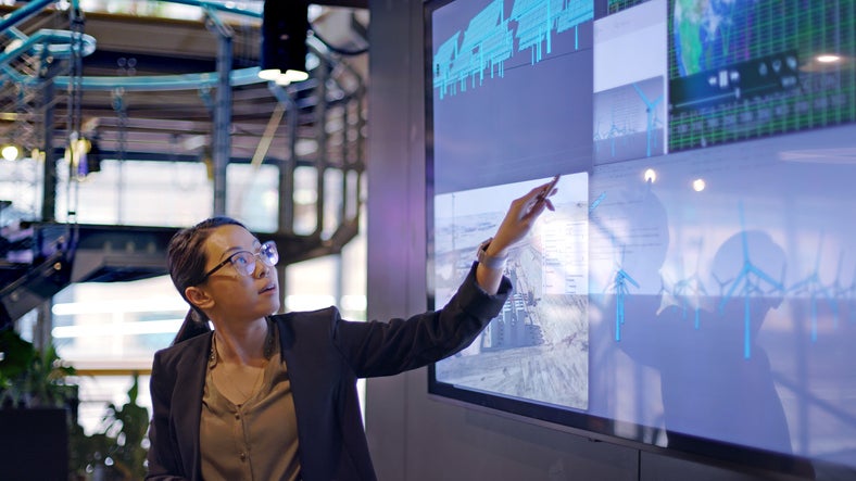 Stock photograph of a young Asian woman conducting a seminar / lecture with the aid of a large screen. The screen is displaying data&designs concerning low carbon electricity production with solar panels&wind turbines. These are juxtaposed with an image of conventional fossil fuel oil production.