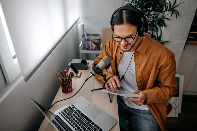 Young male podcaster recording audio using laptop and headset holding his notes and talking on the microphone.