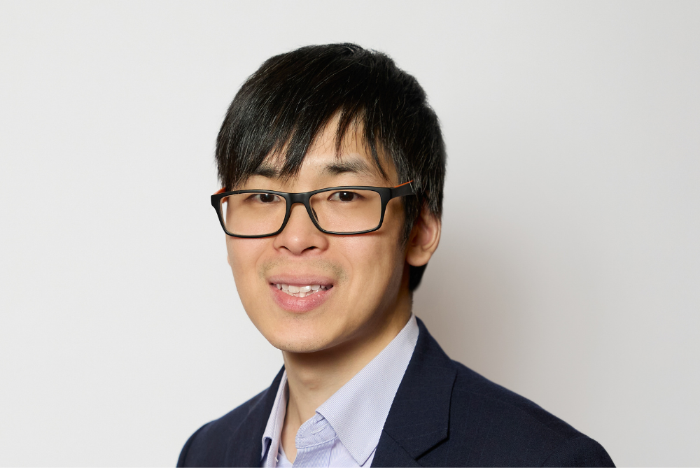 Business man smiling at camera with black hair, black rimmed glasses, and open shirt and black suit