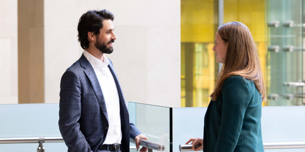 Man in dark business suit leaning in bannister talking to women in a green jacket
