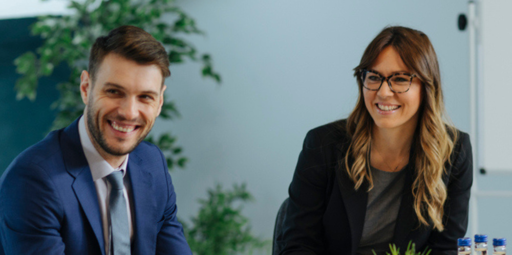 Male in blue jacket and female in black jacket and long brown hair looking off camera around boardroom table