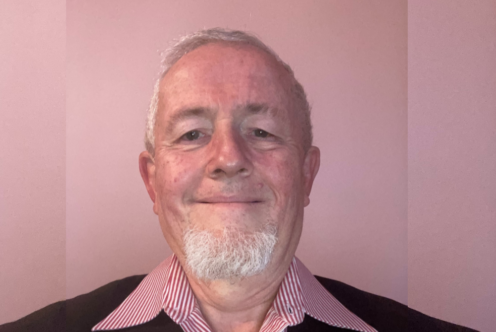 Man wit grey hair and beard smiling at camera, close up photo with background of pink hues.
