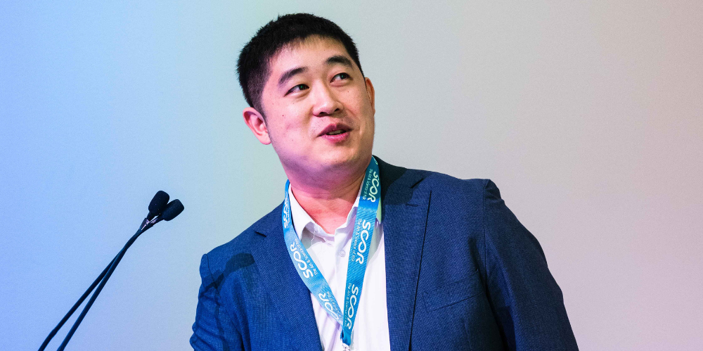 Man at podium looking up and speaking, in blue jacket and white shirt