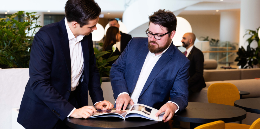 Two professional men stand looking at magazine on a table with people in discussion in background.