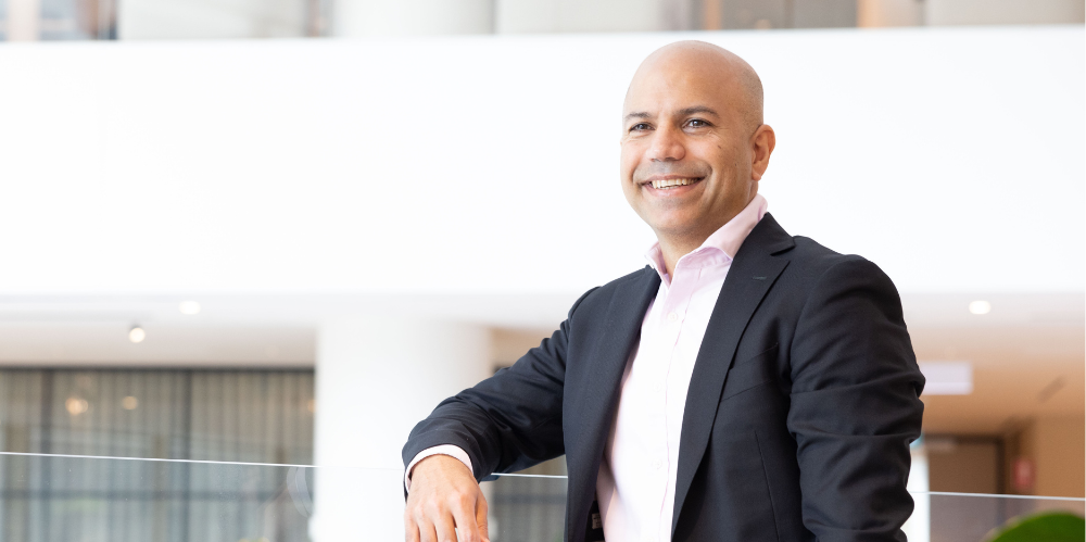 Man standing and leaning on glass bannister smiling looking at camera, in business suit