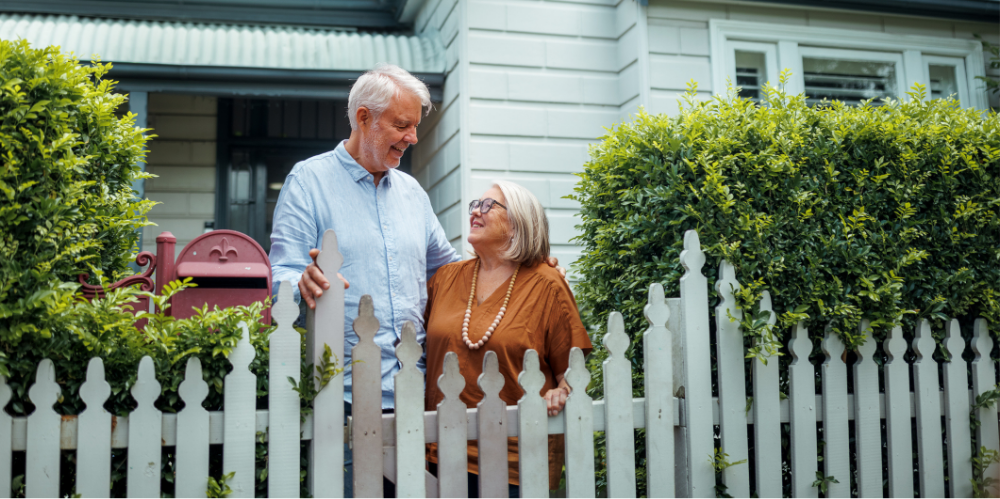 Senior couple embracing while standing near fence in their front yard.