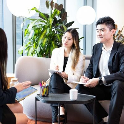 Two young business people sitting on a lounge looking to another woman and chatting. Woman has a white jacket and long black hari and the man has a black suit and open white shirt, holding a notepad and pen.