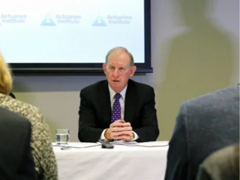 A man in a suit addresses a small audience at a table, with Actuaries Institute branding displayed on a screen behind him.