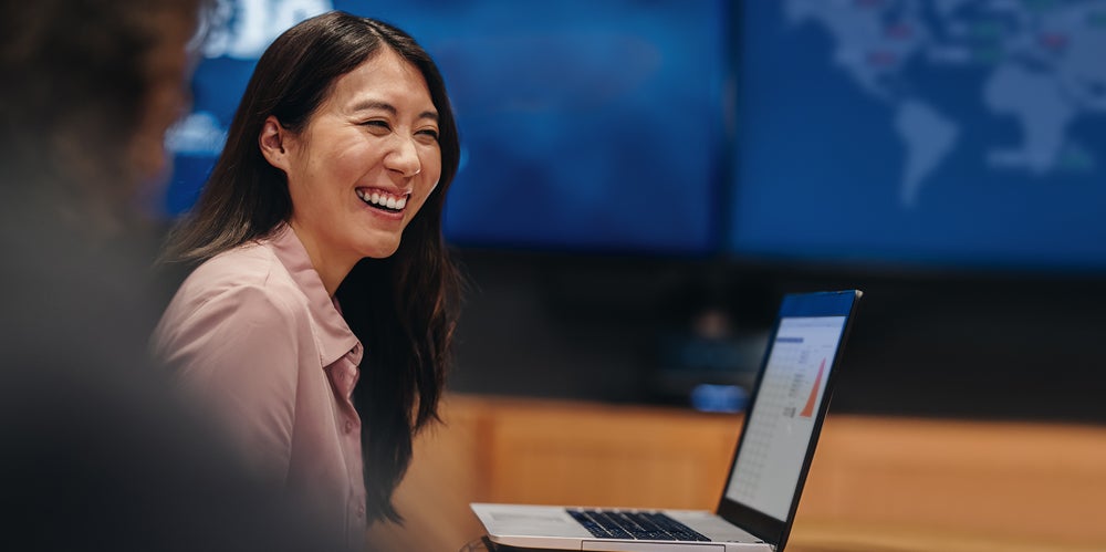 Lady with black hair smiling infront of globe map with laptop open in front of her.