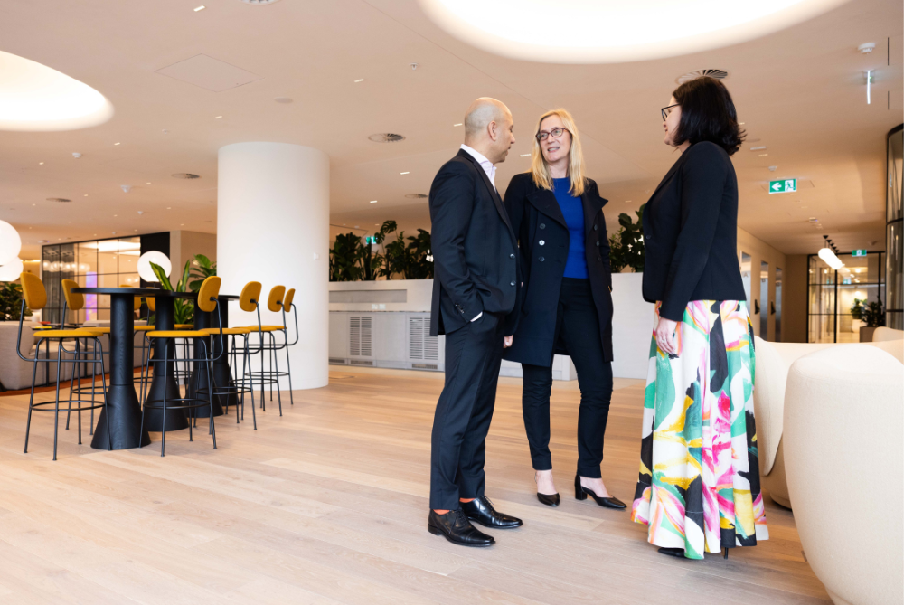 Three business people standing in an office lobby area talking. Left male, and on right 2 females on with a long floral dress and navy jacket