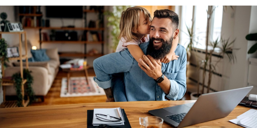 Father working on laptop, with daughter giving him a hug