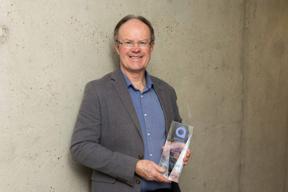 Man with grey jacket and open neck blue shirt, holding tropy and smiling at camera