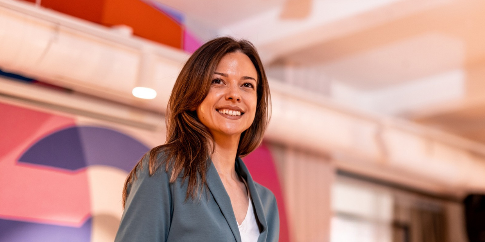 Business women smiligng with light grey jackey on and white blouse, standing in an office space which has pink and lilac hues