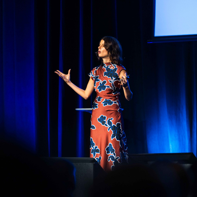 Alix Pearce presenting on stage at the 2026 All Actuaries Summit, speaking with hands raised against a blue-lit backdrop.