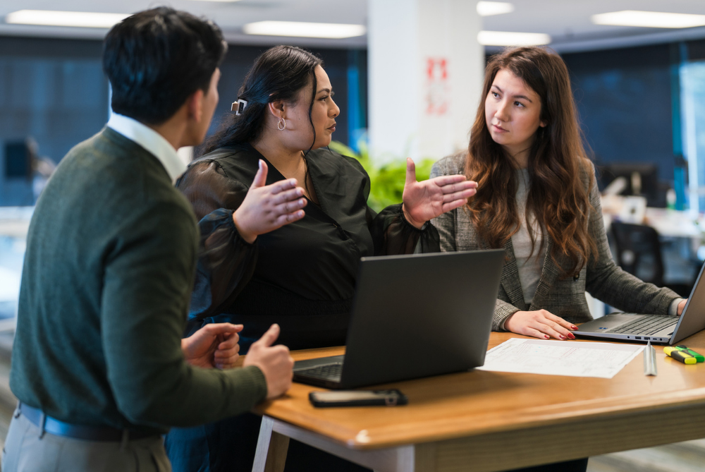 Three people standing at a desk with laptops in a meeting.