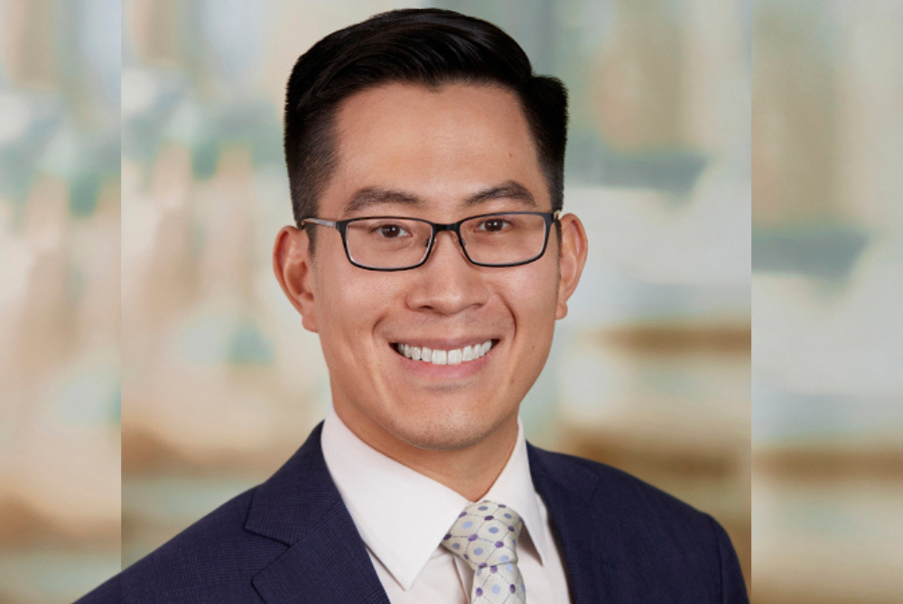 Business man with black hair, black rimmed glasses. Black suit, white shirt and grey and brown checked tie.