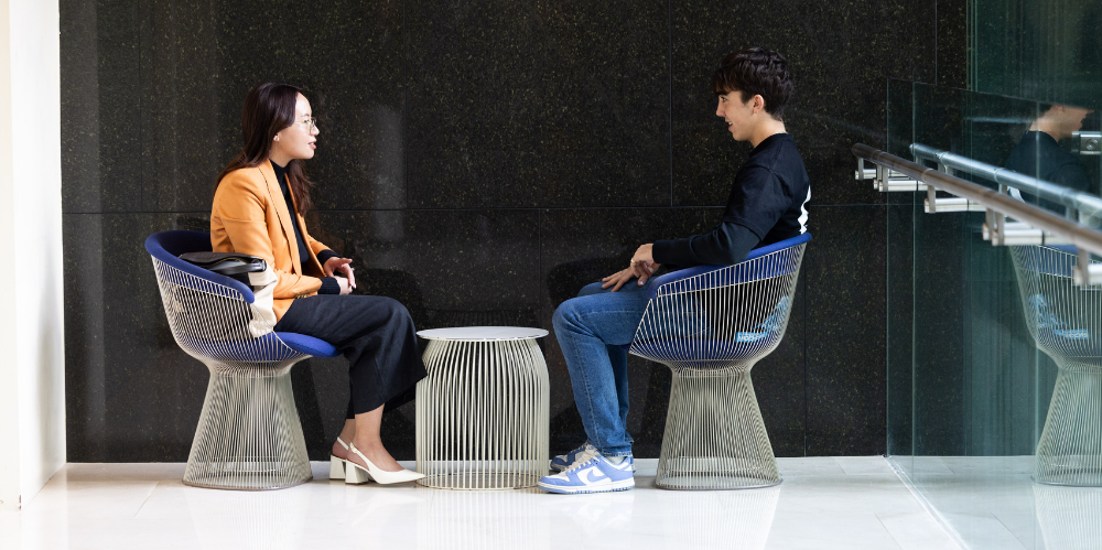TWO people facing eachother, sitting on chair in a foyer and chatting, leaning forward