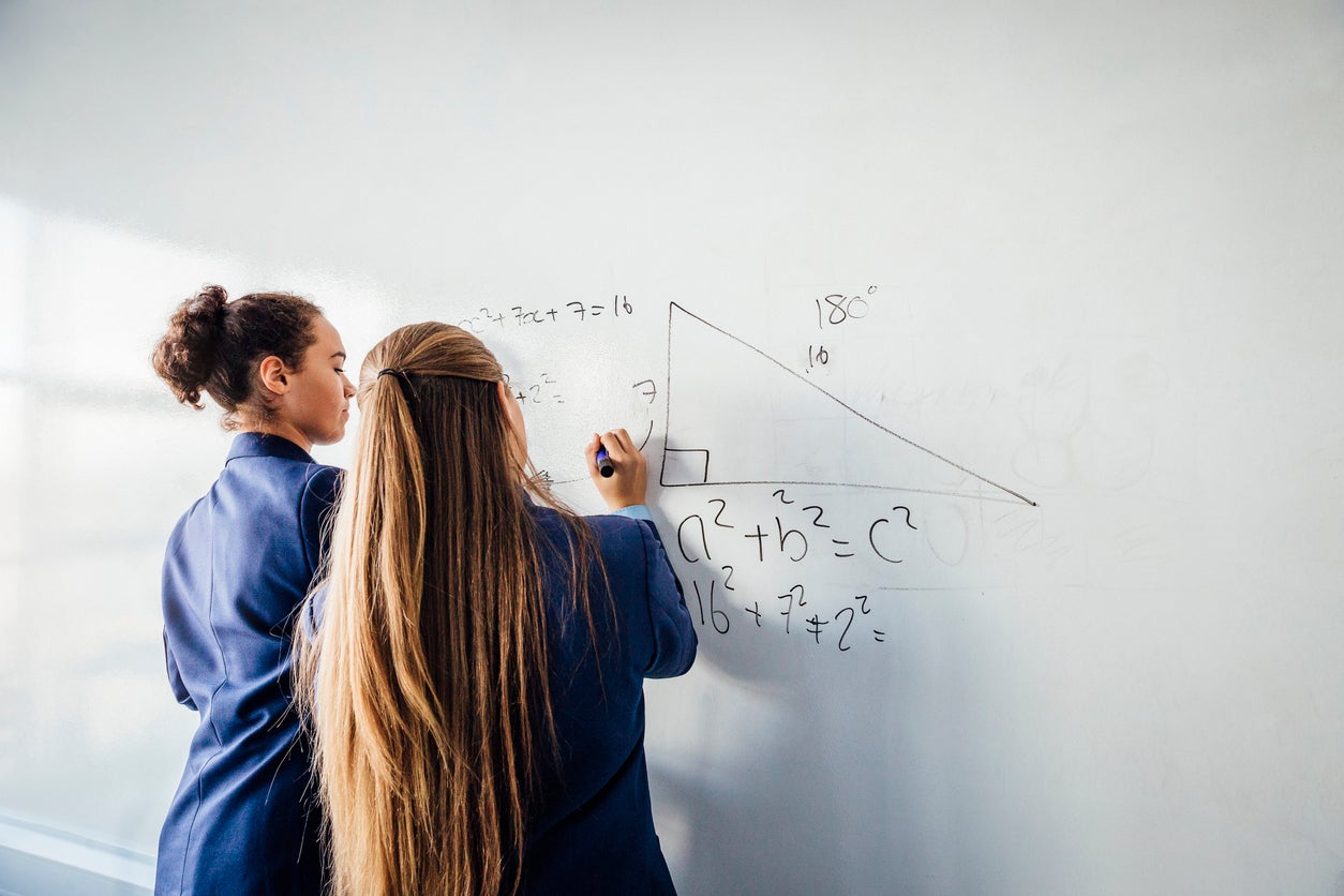 Two teenage school girls standing in front of a large whiteboard side by side solving a mathematics equation on the board. Back view