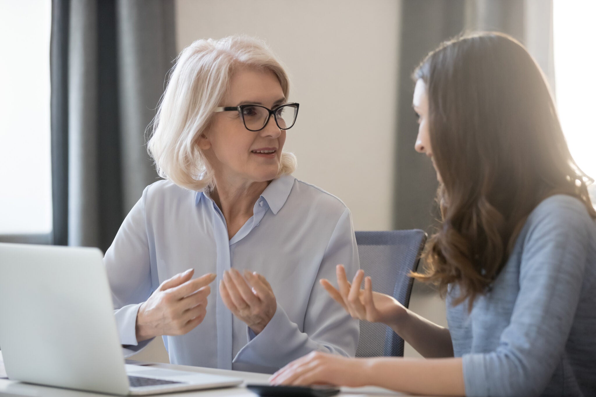 Mature and young women colleagues sitting at desk talking about project startup ideas, sharing thoughts, solve currents issues, make research, discuss growth strategy, think how generate more revenue