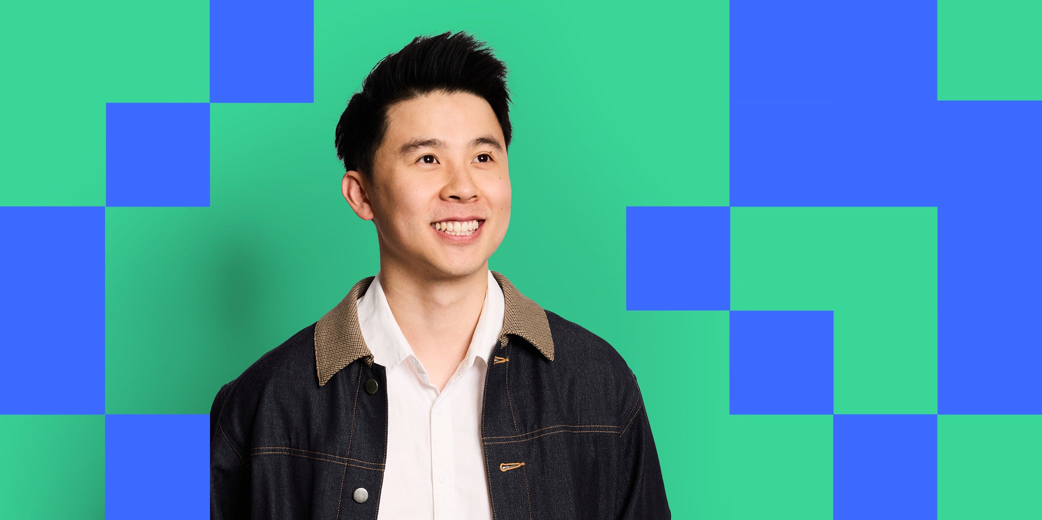 Guy with a big smile looking upwards, has black hair, with dark navy denim jacket and white shirt, on bright blue and green checkered background