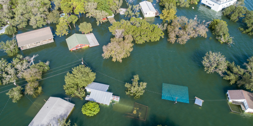 Aerial view of a residential neighbourhood submerged in floodwater, with rooftops, trees, and power lines partially visible above the surface.