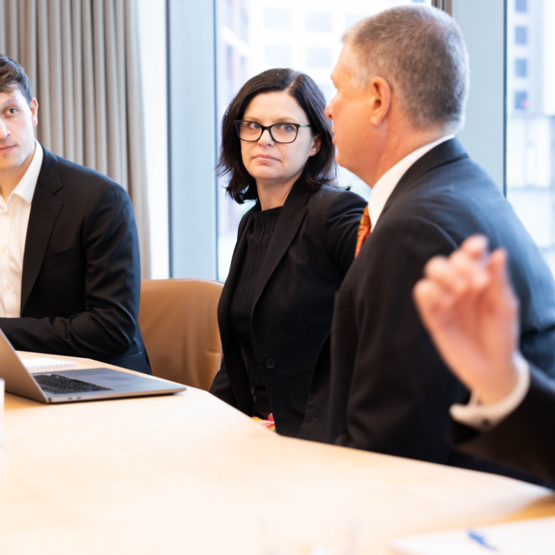 Business people in a meeting, women in the centre with black jacket and glasses looking at a man in suit talking