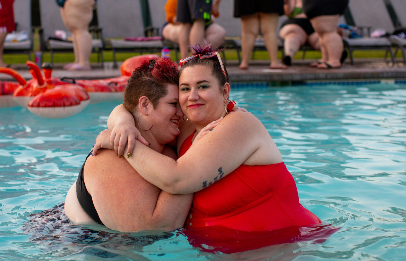 A mid-shot of 2 women, a couple. They are both dressed in swimwear, and are standing waist-deep in a pool. They are embracing, one is laughing and one is smiling. 