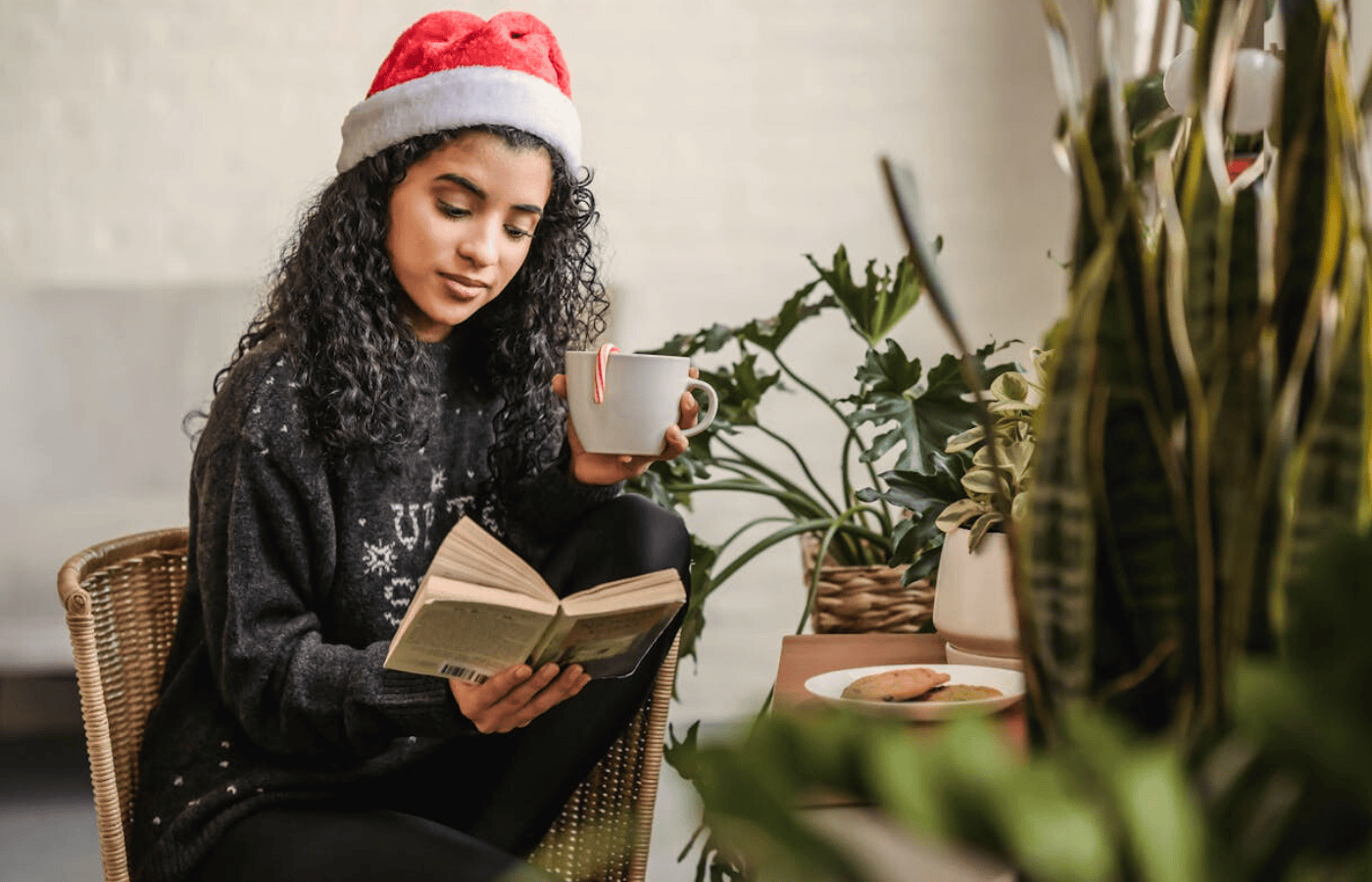 A mid-shot of a woman in a Father Christmas/Santa Claus hat. She drinks a cup of tea and reads a book. 